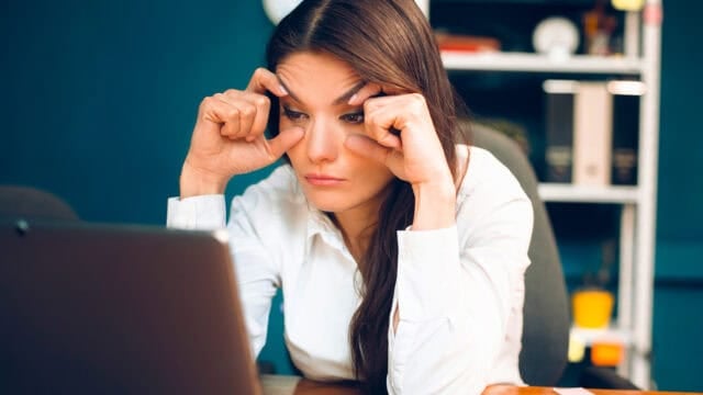 A woman in a white shirt sits at a desk, looking tired as she rubs her eyes while staring at a laptop in an office setting—a scene that reflects the common traits of procrastination often found among original minds.