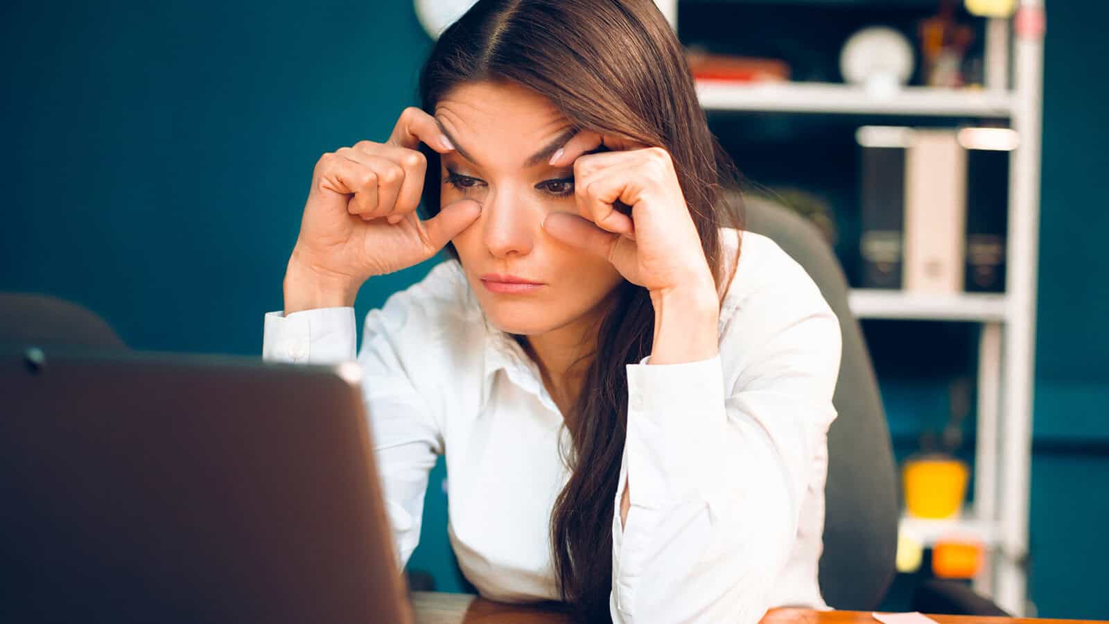 A woman in a white shirt sits at a desk, looking tired as she rubs her eyes while staring at a laptop in an office setting—a scene that reflects the common traits of procrastination often found among original minds.