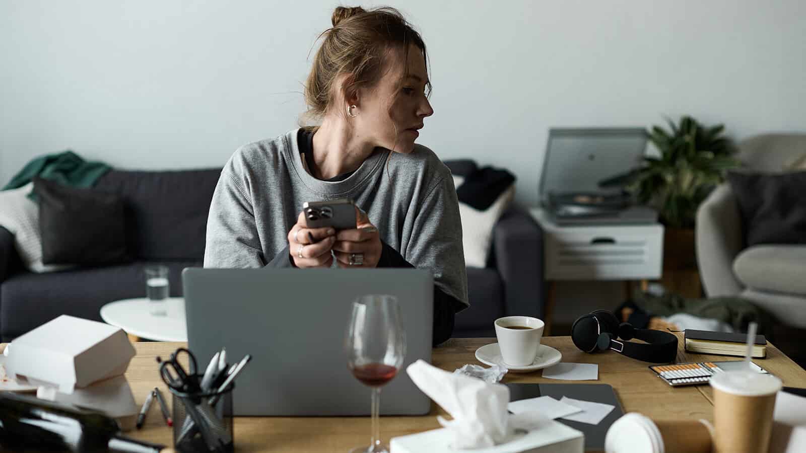 A woman sits at a cluttered table with a laptop, holding a phone and looking to the side. Amidst the messy desks atmosphere, coffee, wine, tissues, and papers are scattered across the surface—a testament to her creative advantage—while a living room provides the backdrop.