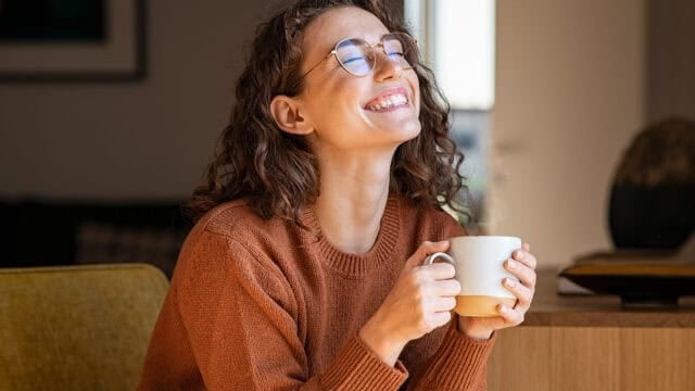 Woman with curly hair and glasses, wearing a brown sweater, sits indoors smiling with eyes closed, holding a steaming white mug in both hands, appearing relaxed and content—an embodiment of comfort often found in minimalist homes.