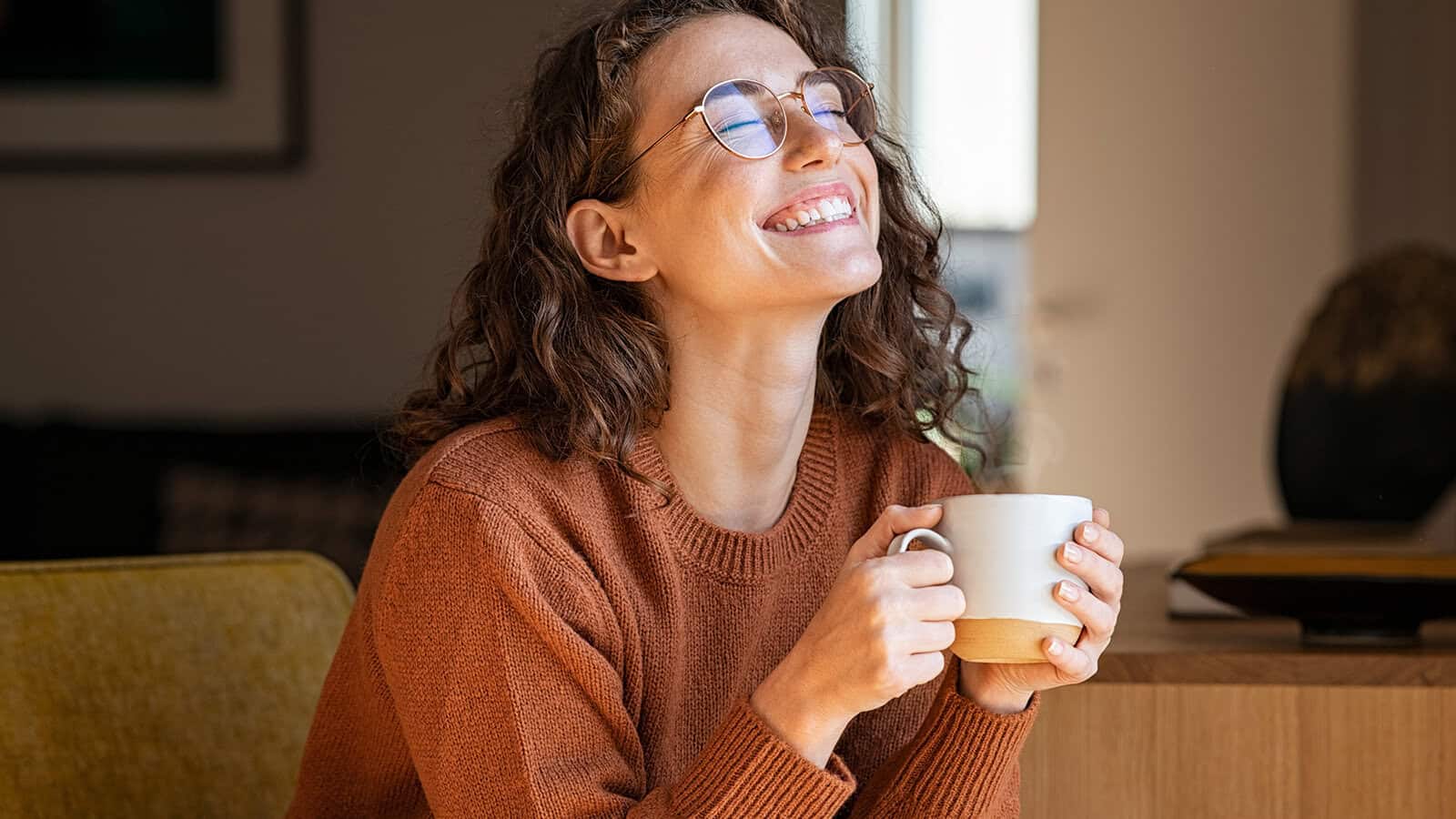 Woman with curly hair and glasses, wearing a brown sweater, sits indoors smiling with eyes closed, holding a steaming white mug in both hands, appearing relaxed and content—an embodiment of comfort often found in minimalist homes.