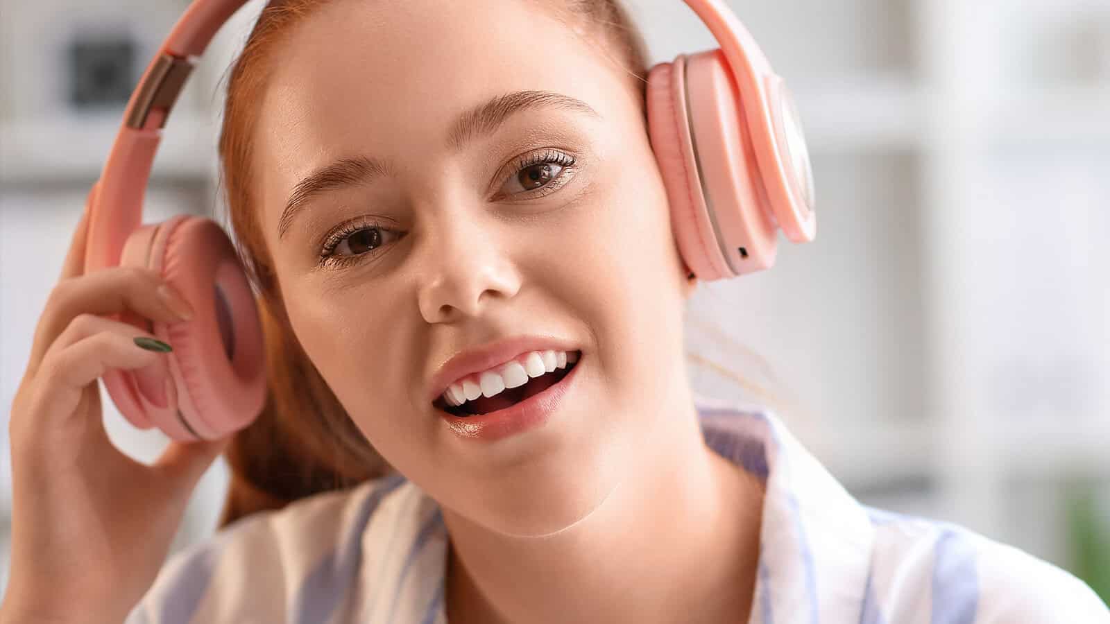 A young person with red hair smiles while wearing pink headphones and a striped shirt, holding one earcup with their hand—perhaps immersed in music with lyrics that help them concentrate. The background is softly blurred.