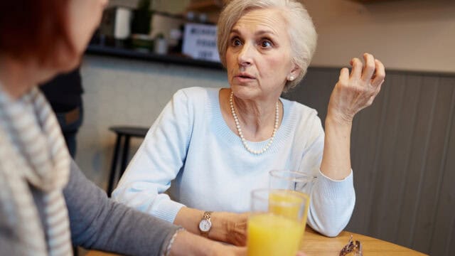 Older woman with short gray hair, wearing a light blue sweater and pearl necklace, sits at a table with glasses of orange juice, appearing engaged in daily conversation with another person whose face is not fully visible. This moment captures the warmth and connection often found in meaningful exchanges during later life.