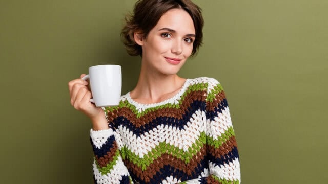 A person with short brown hair, wearing a colorful zigzag-patterned sweater, holds a white mug of coffee black and smiles slightly while standing in front of a plain olive green background.