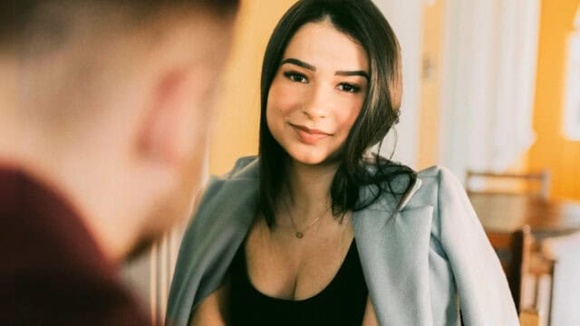 A woman with long dark hair and a light blue jacket smiles at someone in the foreground, who is out of focus, capturing a fleeting moment of social interaction. The background is softly lit with warm tones.