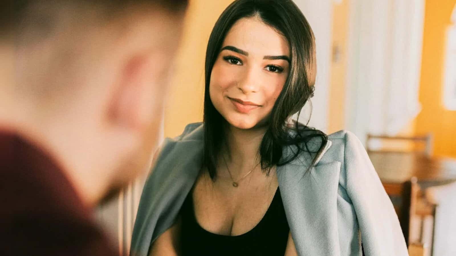 A woman with long dark hair and a light blue jacket smiles at someone in the foreground, who is out of focus, capturing a fleeting moment of social interaction. The background is softly lit with warm tones.