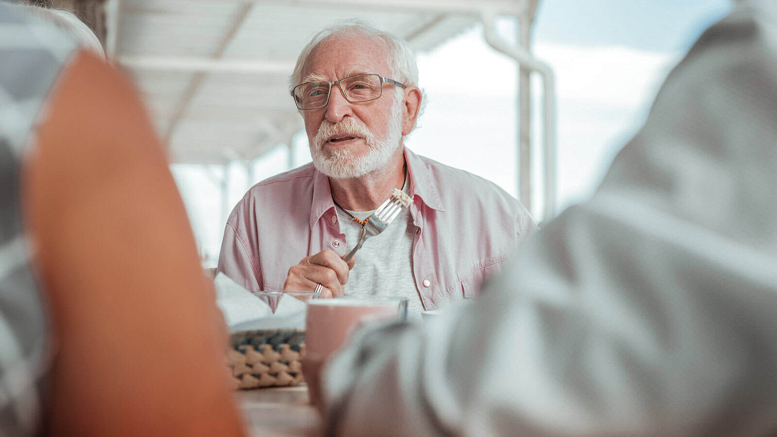 An older man with white hair and glasses, likely from the boomer generation, wearing a light pink shirt, sits at a table holding a fork, engaged in conversation with others at an outdoor setting.