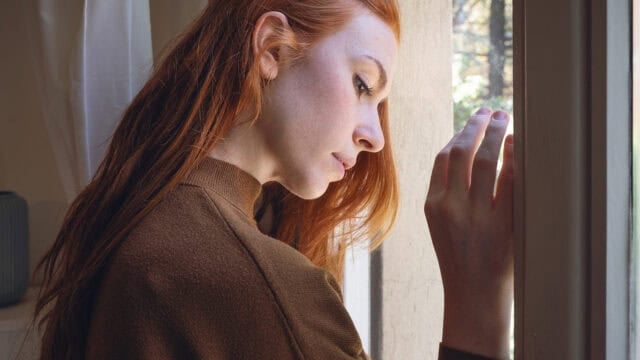 A generous person with long red hair and a brown sweater looks pensively out of a window, resting her hand on the glass with natural light illuminating her face.