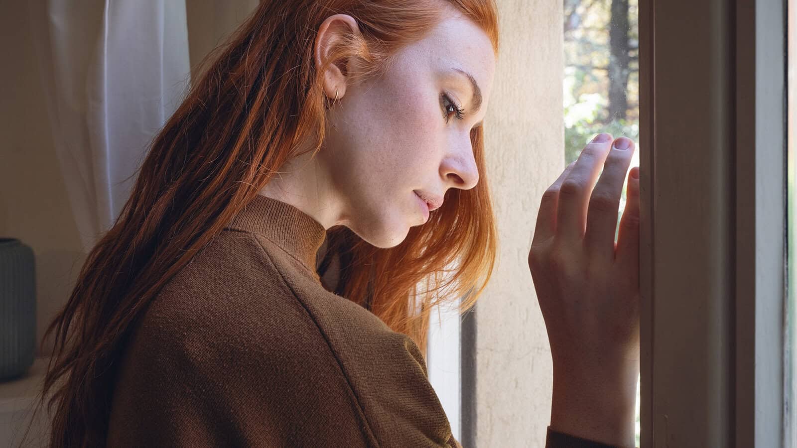 A generous person with long red hair and a brown sweater looks pensively out of a window, resting her hand on the glass with natural light illuminating her face.