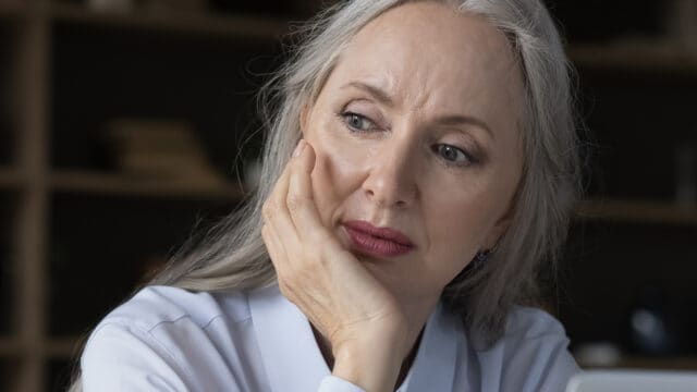 Older woman with long gray hair rests her face on her hand, looking thoughtfully to the side. She wears a white shirt and sits indoors with shelves blurred in the background, reflecting a psychology concept known as socioemotional selectivity, often marked by a shrinking social circle in later life.