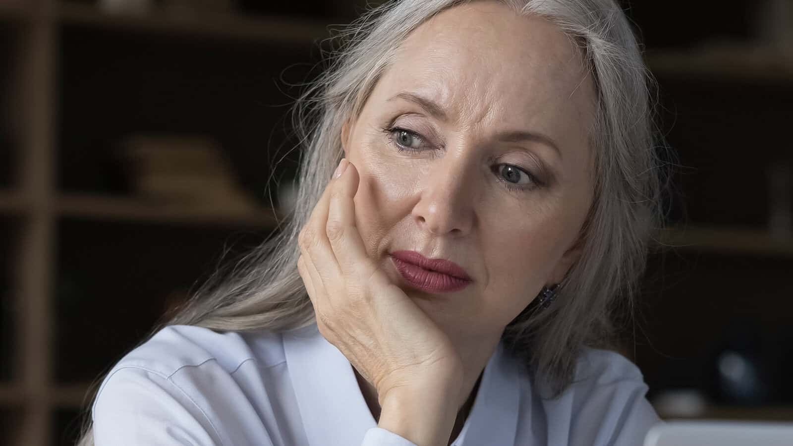 Older woman with long gray hair rests her face on her hand, looking thoughtfully to the side. She wears a white shirt and sits indoors with shelves blurred in the background, reflecting a psychology concept known as socioemotional selectivity, often marked by a shrinking social circle in later life.