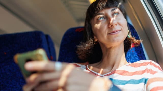 A woman with short dark hair sits by a train window, smiling and looking outside while holding a smartphone. Sunlight shines on her face, and she wears a striped shirt and hoop earrings, perhaps reflecting on travel memories as she savors the journey according to the psychology of travel.