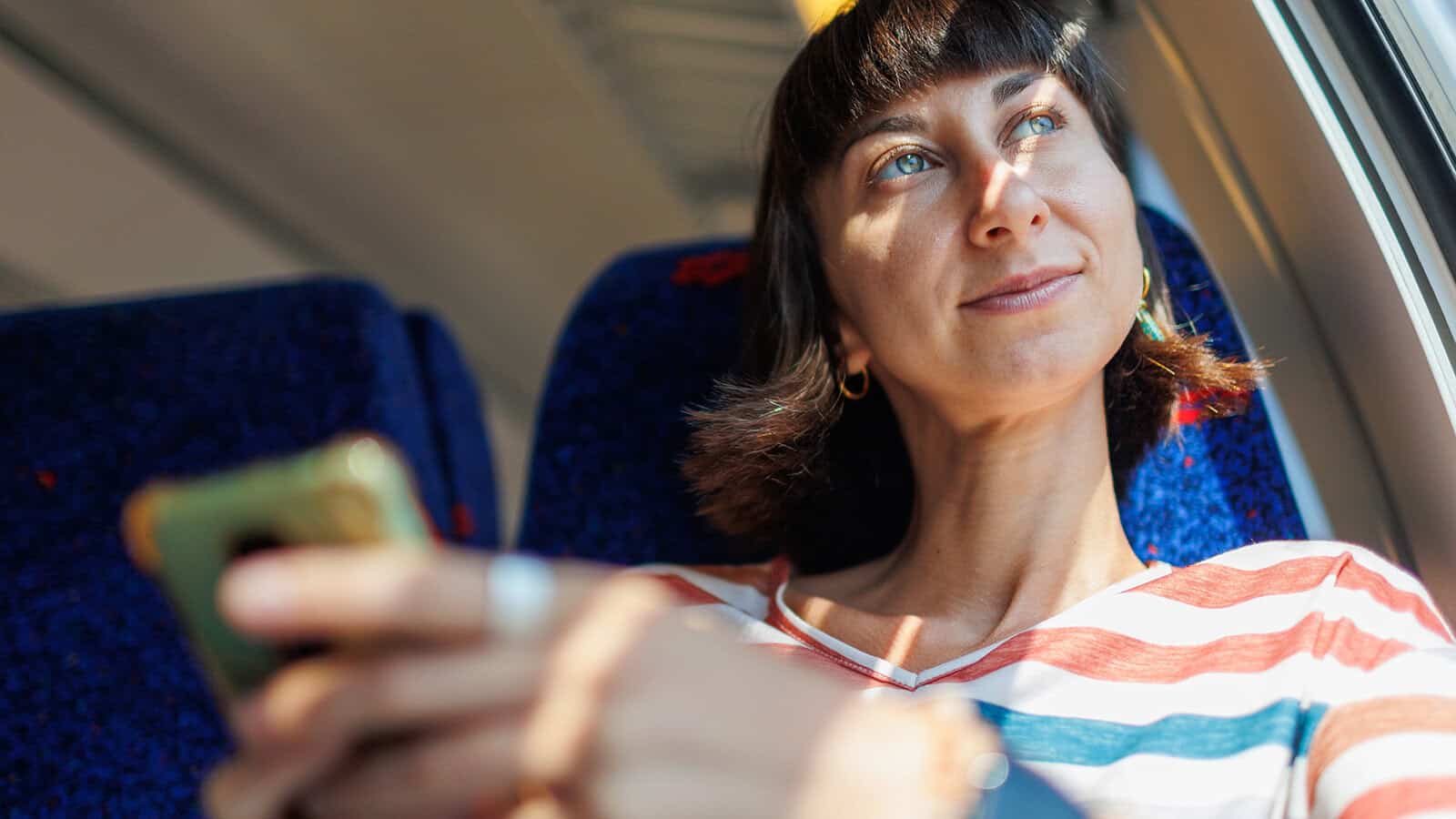 A woman with short dark hair sits by a train window, smiling and looking outside while holding a smartphone. Sunlight shines on her face, and she wears a striped shirt and hoop earrings, perhaps reflecting on travel memories as she savors the journey according to the psychology of travel.