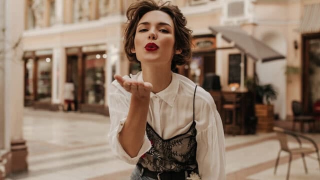 A woman with short brown hair and red lipstick blows a kiss toward the camera while standing in a stylish, outdoor shopping area lined with shops and cafes, capturing a playful moment that hints at confidence and positive self-perception.