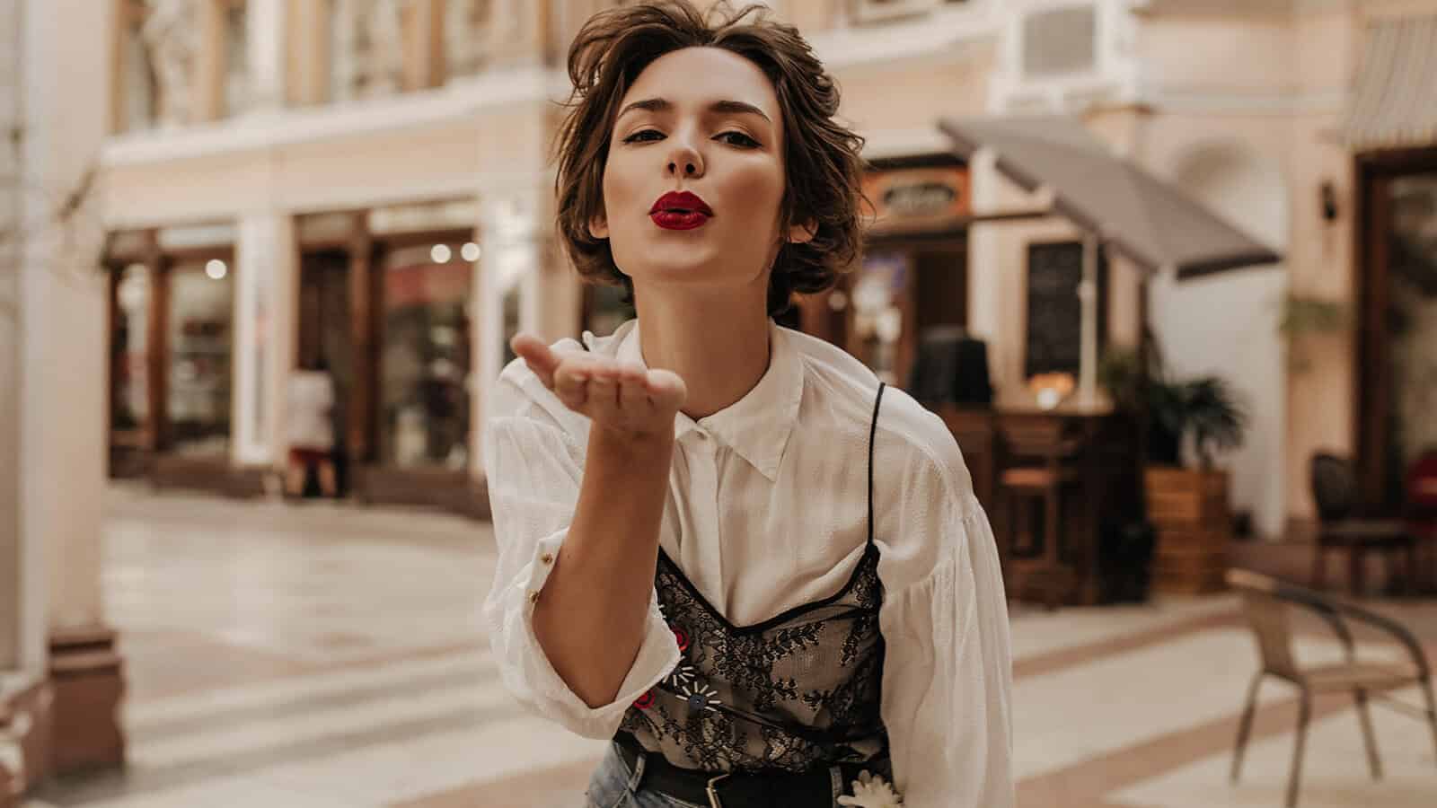A woman with short brown hair and red lipstick blows a kiss toward the camera while standing in a stylish, outdoor shopping area lined with shops and cafes, capturing a playful moment that hints at confidence and positive self-perception.