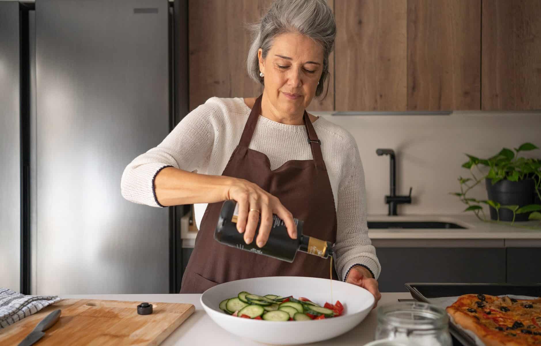 A woman in a brown apron pours olive oil onto a fresh salad in a kitchen. A wooden cutting board and a tray of food are on the counter nearby.