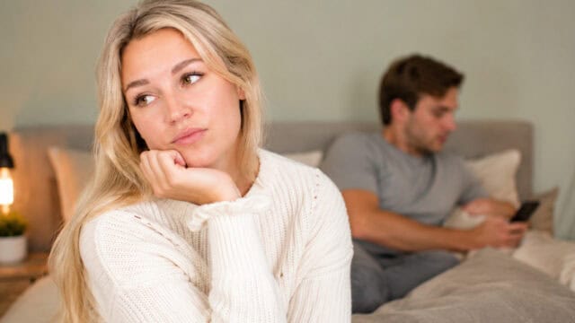 A woman in a white sweater looks sad and thoughtful while sitting on a bed, with a man in the background looking at his phone, both appearing distant—a scene that may reflect subtle ex-partner signs and is often discussed in relationship advice about recognizing when your partner isn’t fully present.