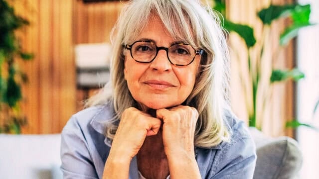 An older woman with gray hair and glasses rests her chin on her hands, smiling gently. She sits indoors in a cozy room with plants and soft lighting in the background.