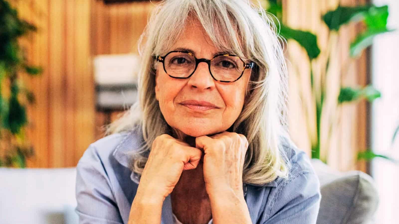 An older woman with gray hair and glasses rests her chin on her hands, smiling gently. She sits indoors in a cozy room with plants and soft lighting in the background.