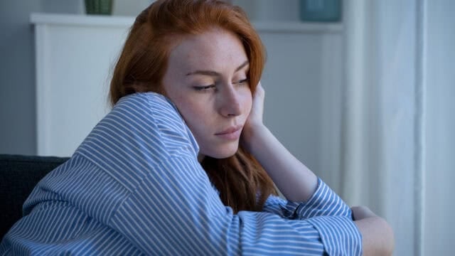 A woman with long red hair wearing a blue striped shirt sits indoors, resting her head on her hand with a thoughtful or sad expression, perhaps reflecting on the loneliness that can come with being the first in your family to achieve something significant.