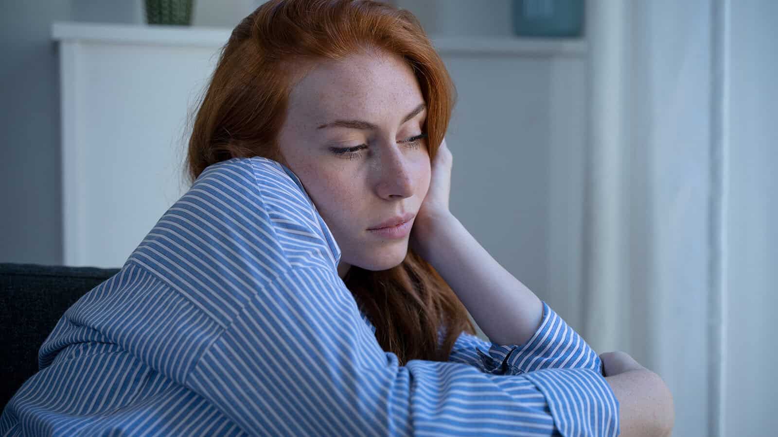 A woman with long red hair wearing a blue striped shirt sits indoors, resting her head on her hand with a thoughtful or sad expression, perhaps reflecting on the loneliness that can come with being the first in your family to achieve something significant.