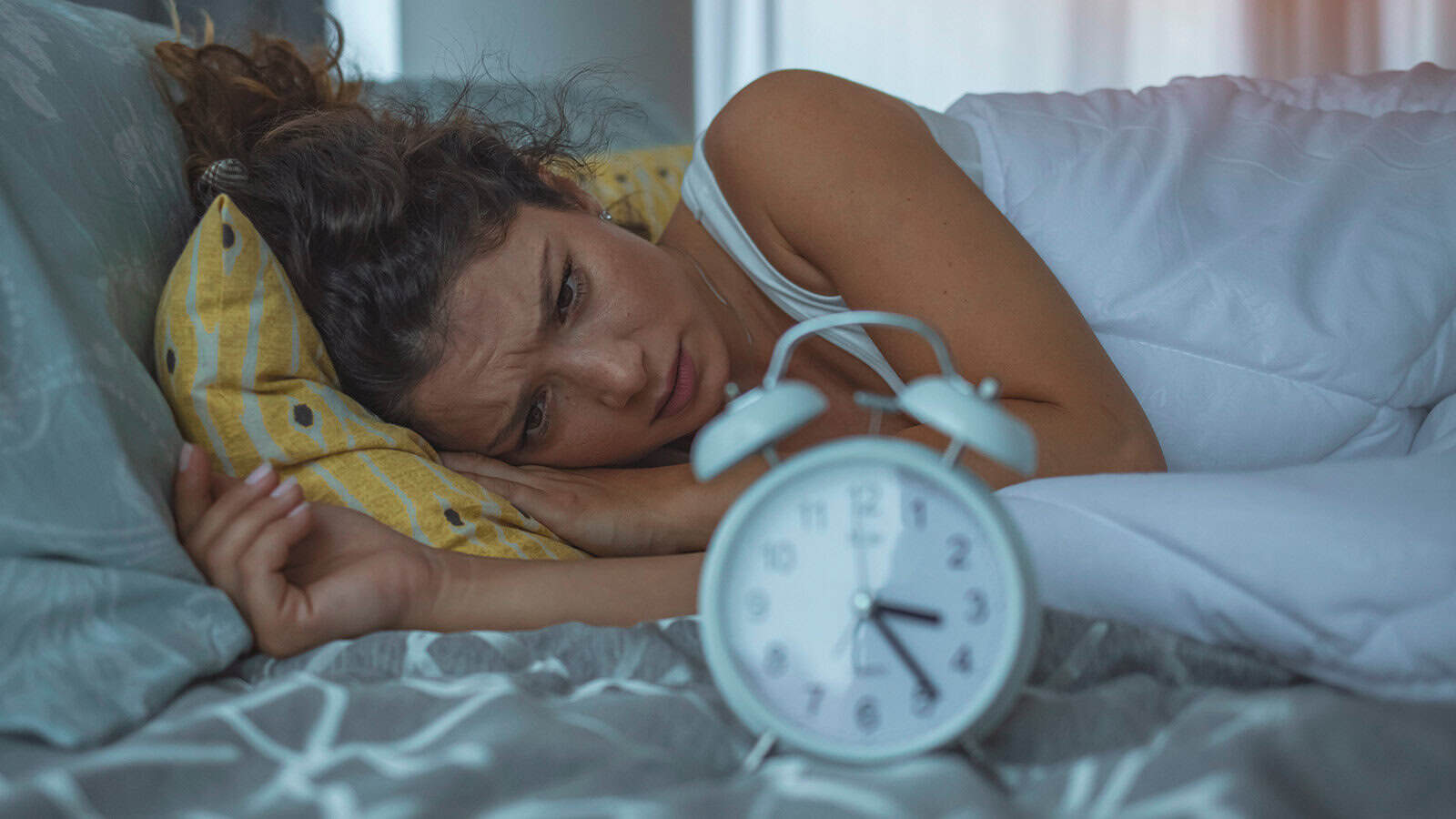 A woman lies in bed awake, looking concerned, her head on a yellow pillow and a white alarm clock showing 5:00 in the foreground, suggesting she is struggling with broken sleep or perhaps facing the familiar 3am problem.