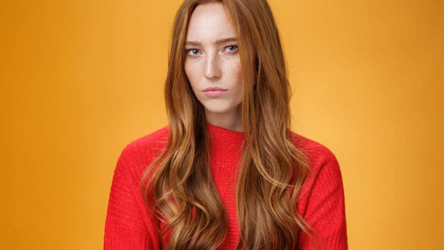 A woman with long red hair wearing a red sweater stands against an orange background, looking directly at the camera with a neutral expression.