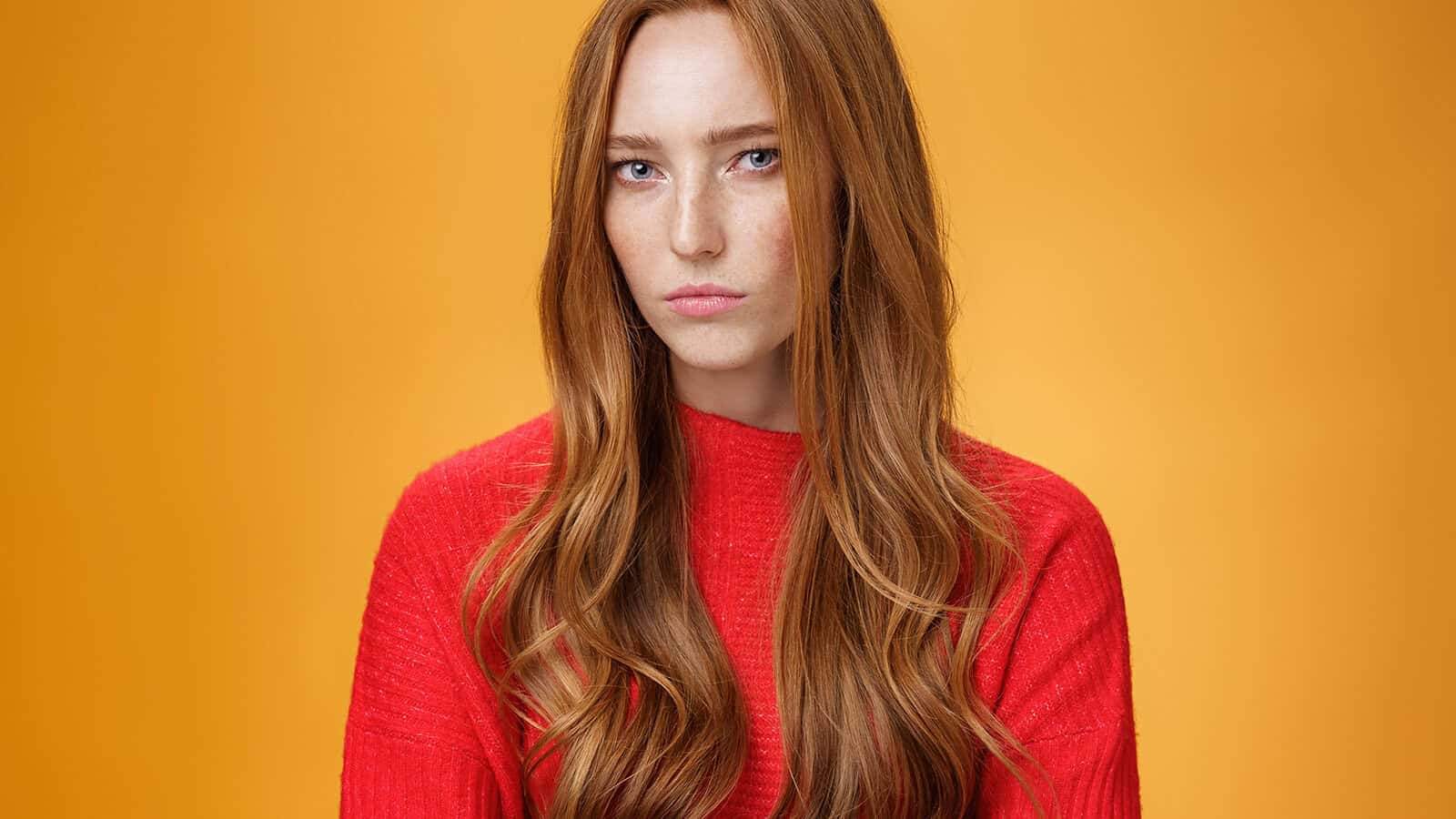 A woman with long red hair wearing a red sweater stands against an orange background, looking directly at the camera with a neutral expression.