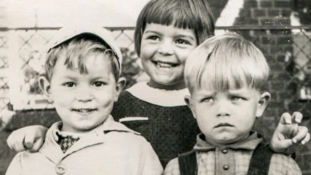 Three young children pose for a vintage black-and-white photo. The girl in the middle smiles brightly, while the boys on either side show different expressions—one smiles, the other looks unhappy. They stand outdoors near a fence.