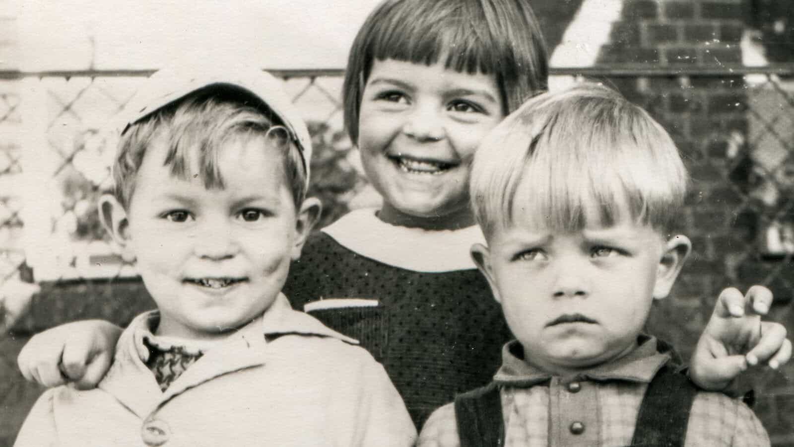Three young children pose for a vintage black-and-white photo. The girl in the middle smiles brightly, while the boys on either side show different expressions—one smiles, the other looks unhappy. They stand outdoors near a fence.