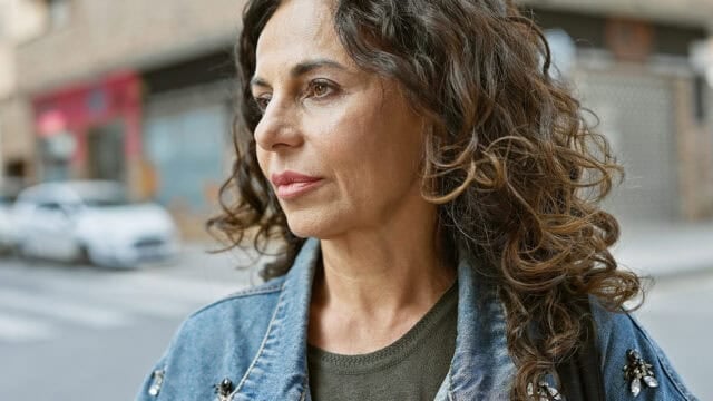 A woman with wavy brown hair wearing a denim jacket stands outdoors on a city street, looking thoughtfully into the distance.
