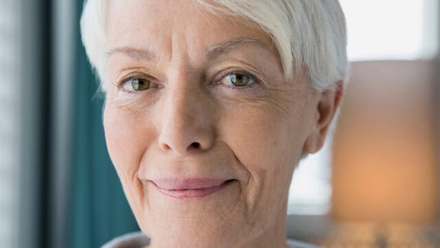 Close-up of an older woman with short white hair, green eyes, and a slight smile, standing indoors with soft natural light illuminating her face.