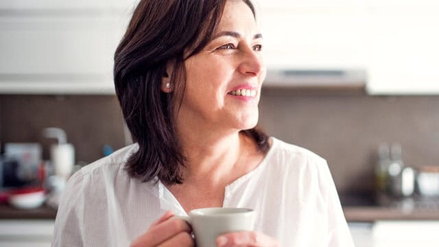 A woman with shoulder-length dark hair, wearing a white shirt, smiles while holding a mug in a bright kitchen.