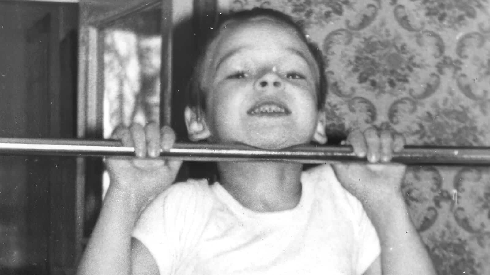 A young child in a white shirt smiles while doing a pull-up on a metal bar indoors, with patterned wallpaper in the background.