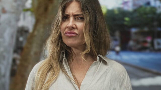 A woman with long, light brown hair wearing a beige shirt stands outdoors, looking skeptical or confused, with her eyebrows furrowed and lips pursed. The background is blurred with trees and sunlight.