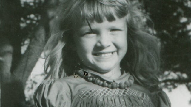 Black and white photo of a young girl with long hair, wearing a beaded necklace and a smocked dress, smiling broadly. Trees are visible in the background.