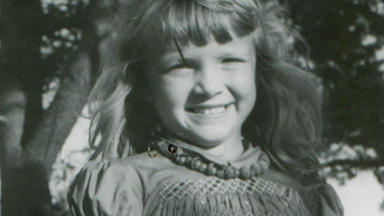 Black and white photo of a young girl with long hair, wearing a beaded necklace and a smocked dress, smiling broadly. Trees are visible in the background.