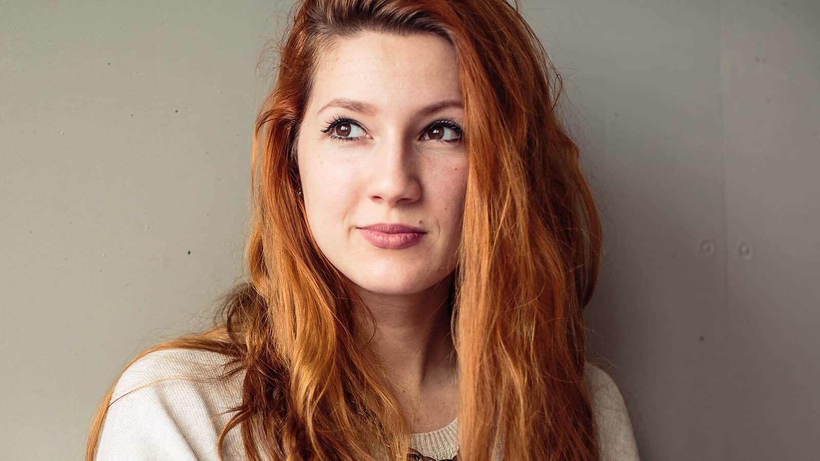 A woman with long red hair and a light sweater looks slightly to the right, standing against a plain, light-colored wall.