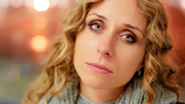 A woman with curly blonde hair and light eyes looks directly at the camera with a serious, slightly sad expression. She is wearing a gray scarf, and the background is softly blurred with warm tones.