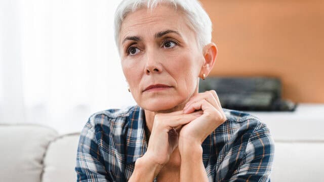 Older woman with short gray hair and a plaid shirt sits indoors, resting her chin on her hands, looking thoughtful and slightly concerned.