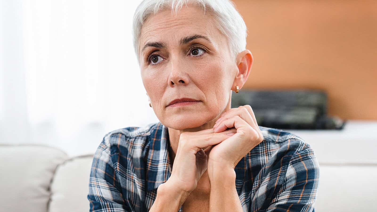 Older woman with short gray hair and a plaid shirt sits indoors, resting her chin on her hands, looking thoughtful and slightly concerned.