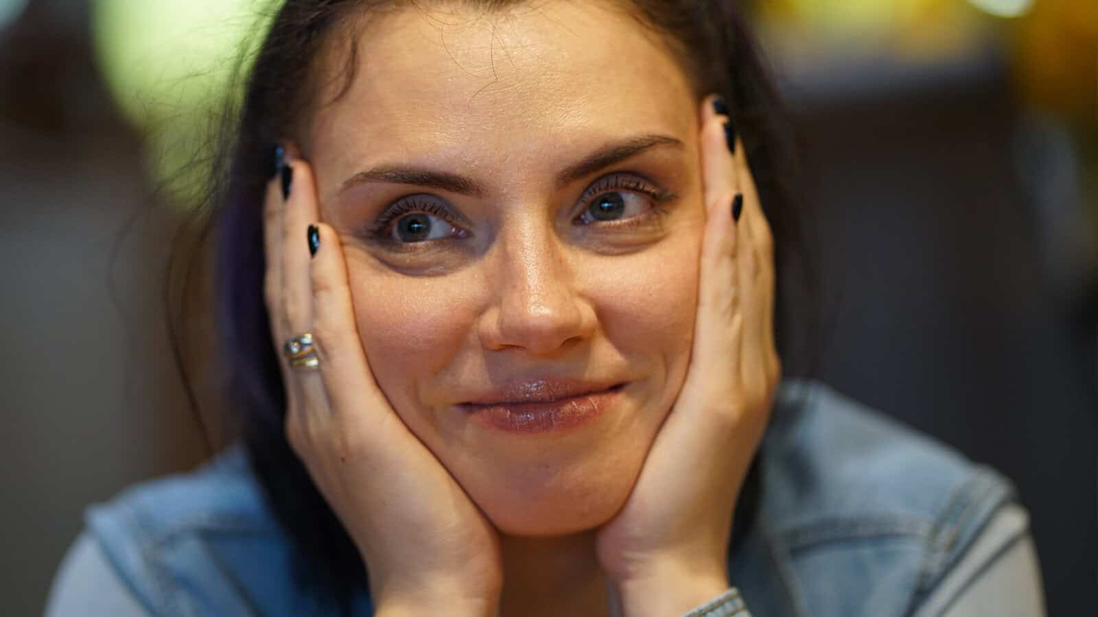 A woman with dark hair smiles softly, resting her face in her hands. She has blue eyes, black nail polish, and wears a denim shirt. The background is blurred with warm lighting.