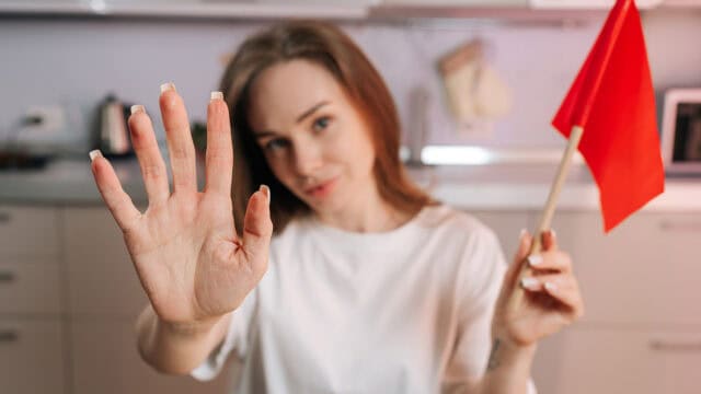 A woman in a white shirt holds up her hand with fingers spread, as if signaling stop, and holds a small red flag in the other hand in a kitchen setting.