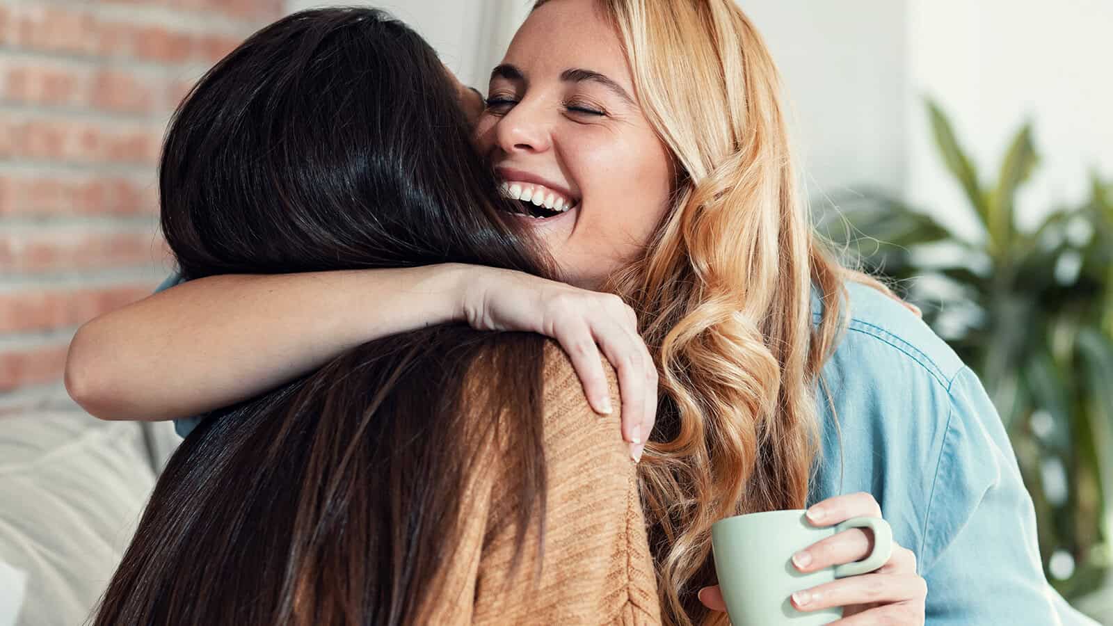 Two women smiling and hugging each other warmly, with one holding a green mug. They appear happy and relaxed, sitting indoors.
