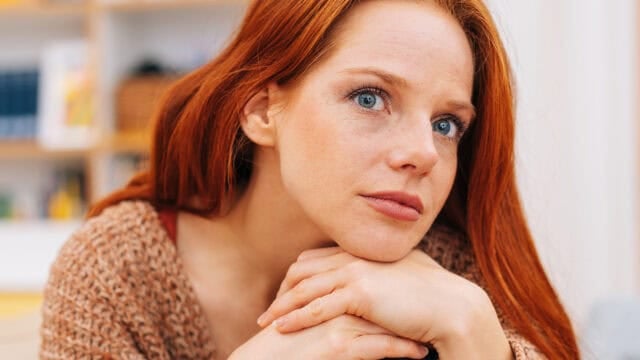 A woman with long red hair and blue eyes rests her chin on her hands and looks thoughtfully at the camera. She is wearing a brown sweater and is indoors, with shelves and blurred background visible.