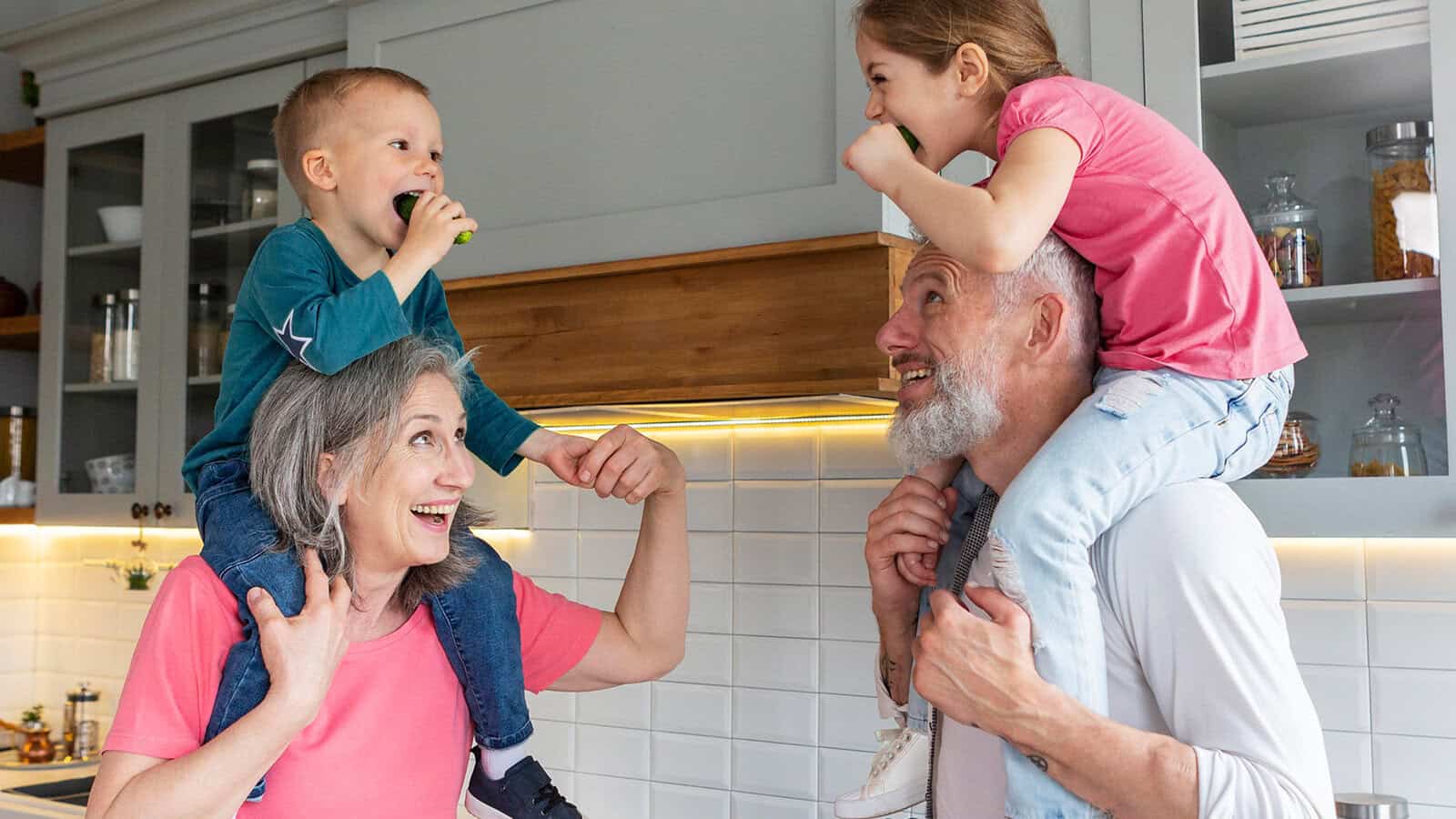 Two happy grandparents give piggyback rides to their smiling grandchildren in a modern kitchen, all laughing and eating vegetables together.