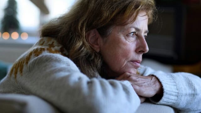 A woman with long brown hair and a patterned sweater rests her chin on her hands, gazing thoughtfully out of a window with soft light illuminating her face.
