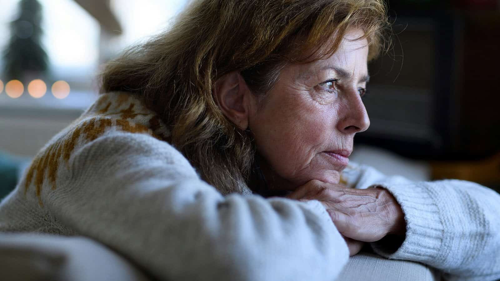 A woman with long brown hair and a patterned sweater rests her chin on her hands, gazing thoughtfully out of a window with soft light illuminating her face.