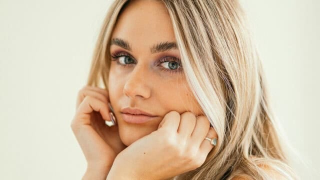 A woman with long blonde hair and light makeup rests her chin on her hands, looking into the camera with a neutral expression. She wears a ring on her finger and has a soft, natural background.