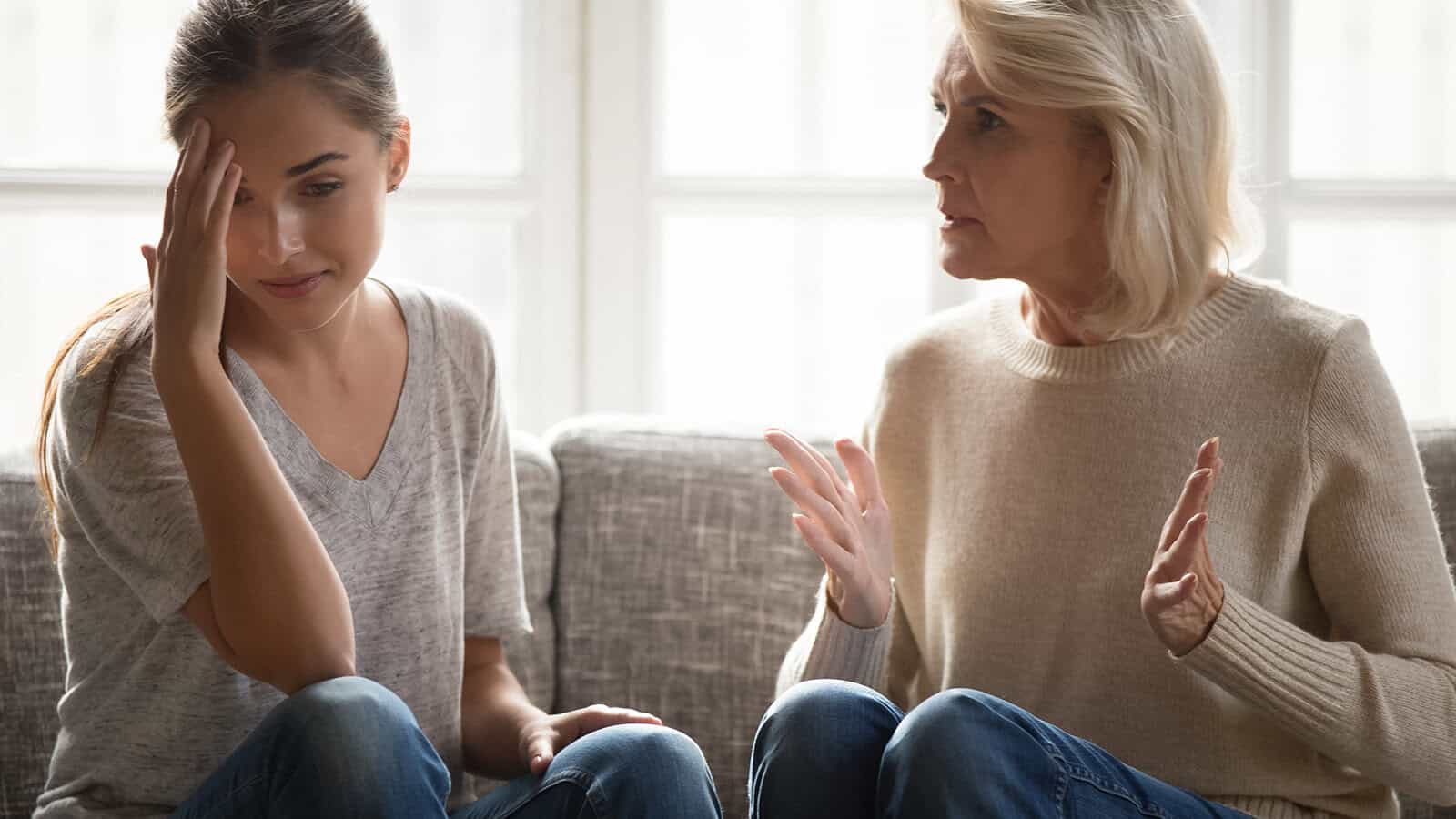 A young woman sits on a couch looking upset with her hand on her forehead, while an older woman next to her speaks with concern, gesturing with her hands.